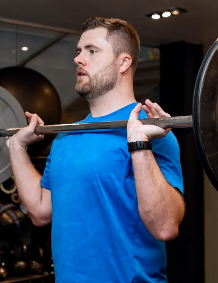 A bearded man holding a barbell across the front of his shoulders in a gym
