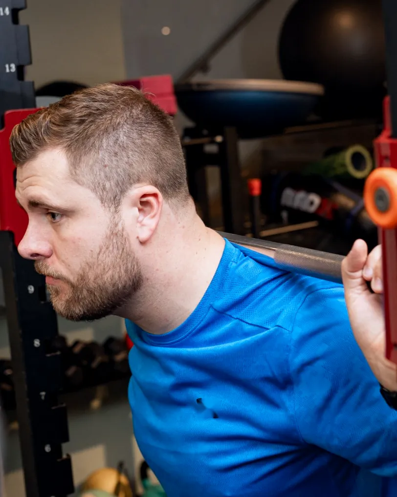 A bearded man in a blue t-shirt in a squat rack with a barbell across his shoulders